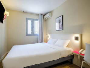 Quarto de hotel com cama, telefone de apoio, unidade de ar condicionado, janela com cortinas e um quadro na parede.