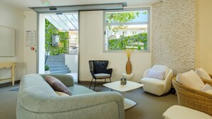 A modern living room with a sofa, armchairs, a coffee table, and a view of a green garden through a large window. - B&B HOTEL Antibes Juan-les-Pins (Antibes)