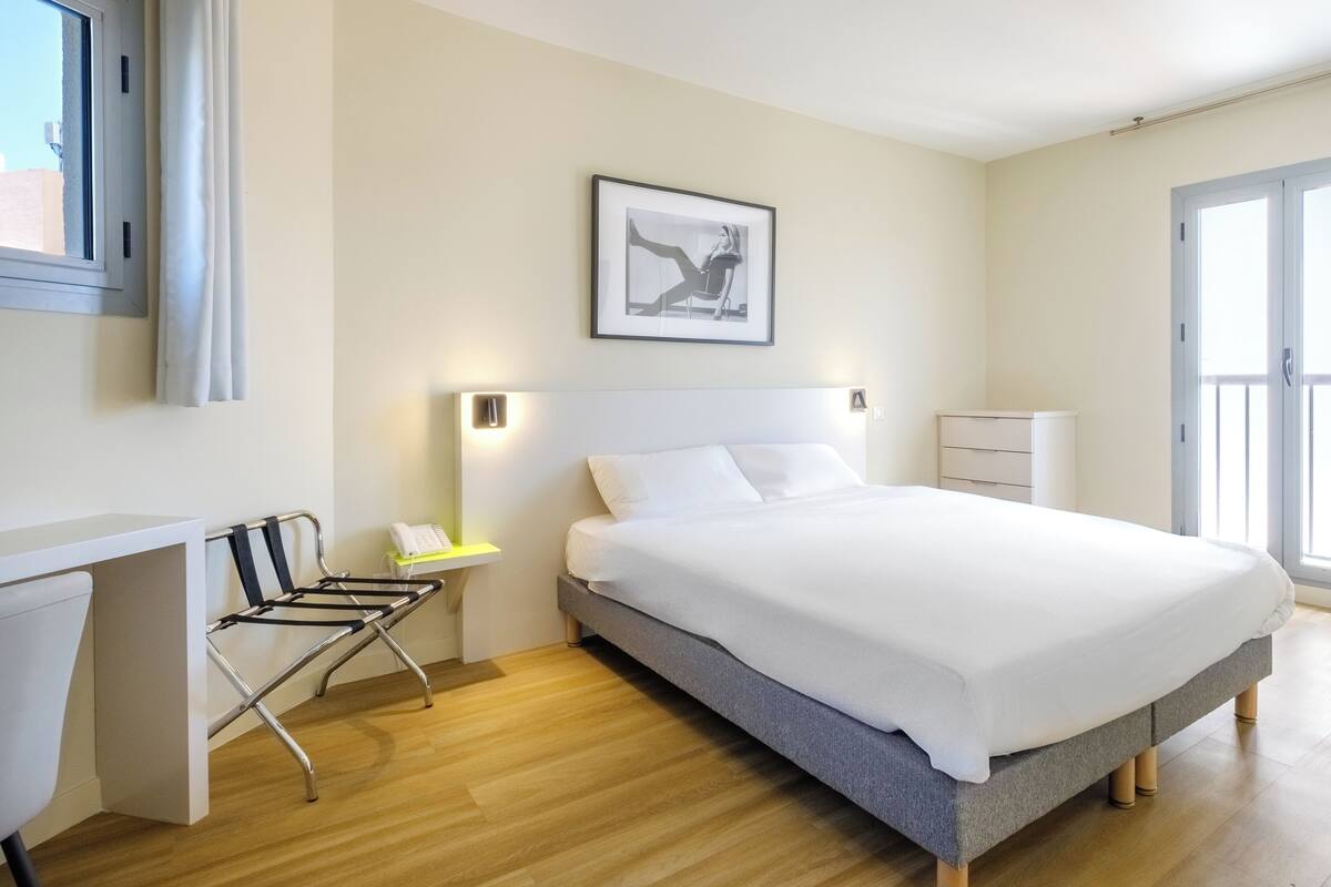 A modern hotel room with a bed, bedside table, lamp, and a desk with a chair.