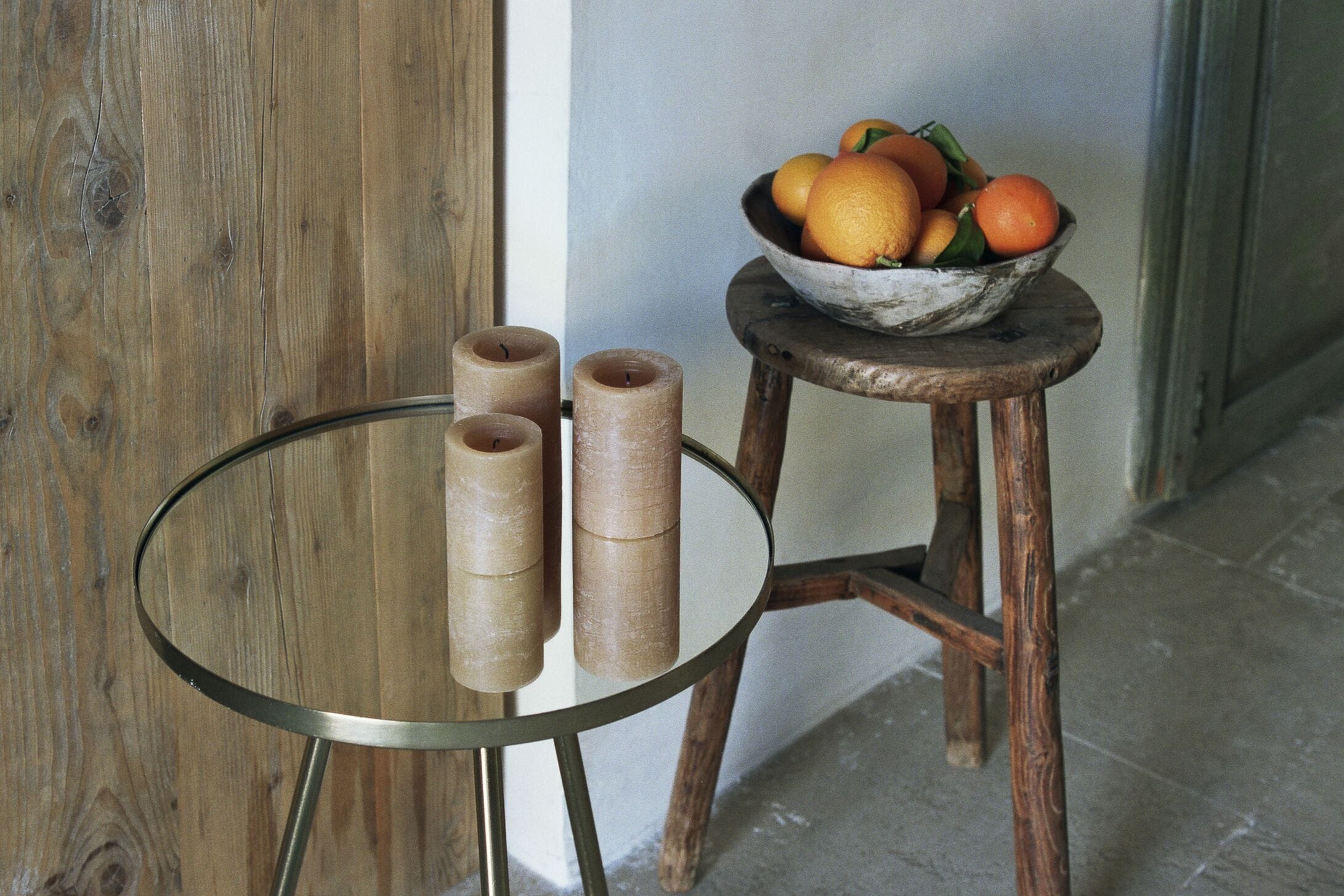 Une table en verre avec trois bougies et un tabouret en bois sur lequel se trouve un bol rempli d’oranges.
