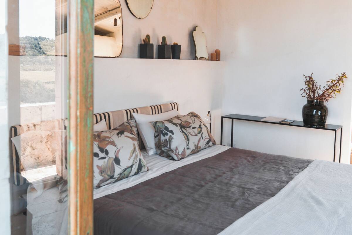 A bedroom with a bed, a bedside table, a mirror, and a vase with flowers.