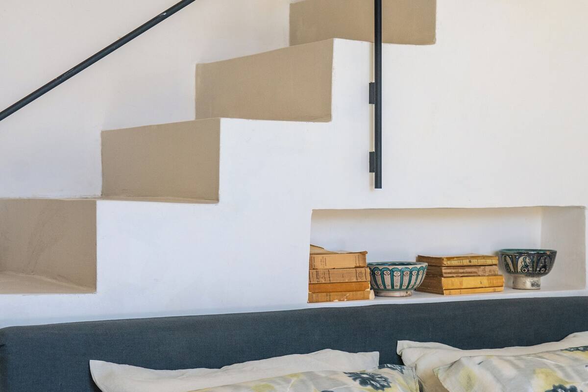 A bedroom with a bed, pillows, a headboard, a staircase, and a shelf with boxes and bowls.