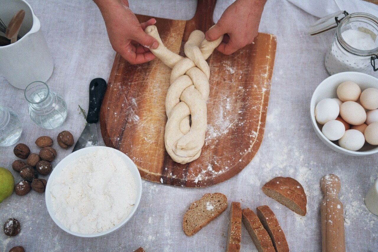 A person braiding dough on a wooden board with flour scattered around, a bowl of flour, a jar of sugar, and a bowl of eggs nearby.