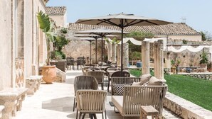Un área de estar al aire libre con mobiliario de mimbre, sombrillas, columnas de piedra y un jardín.