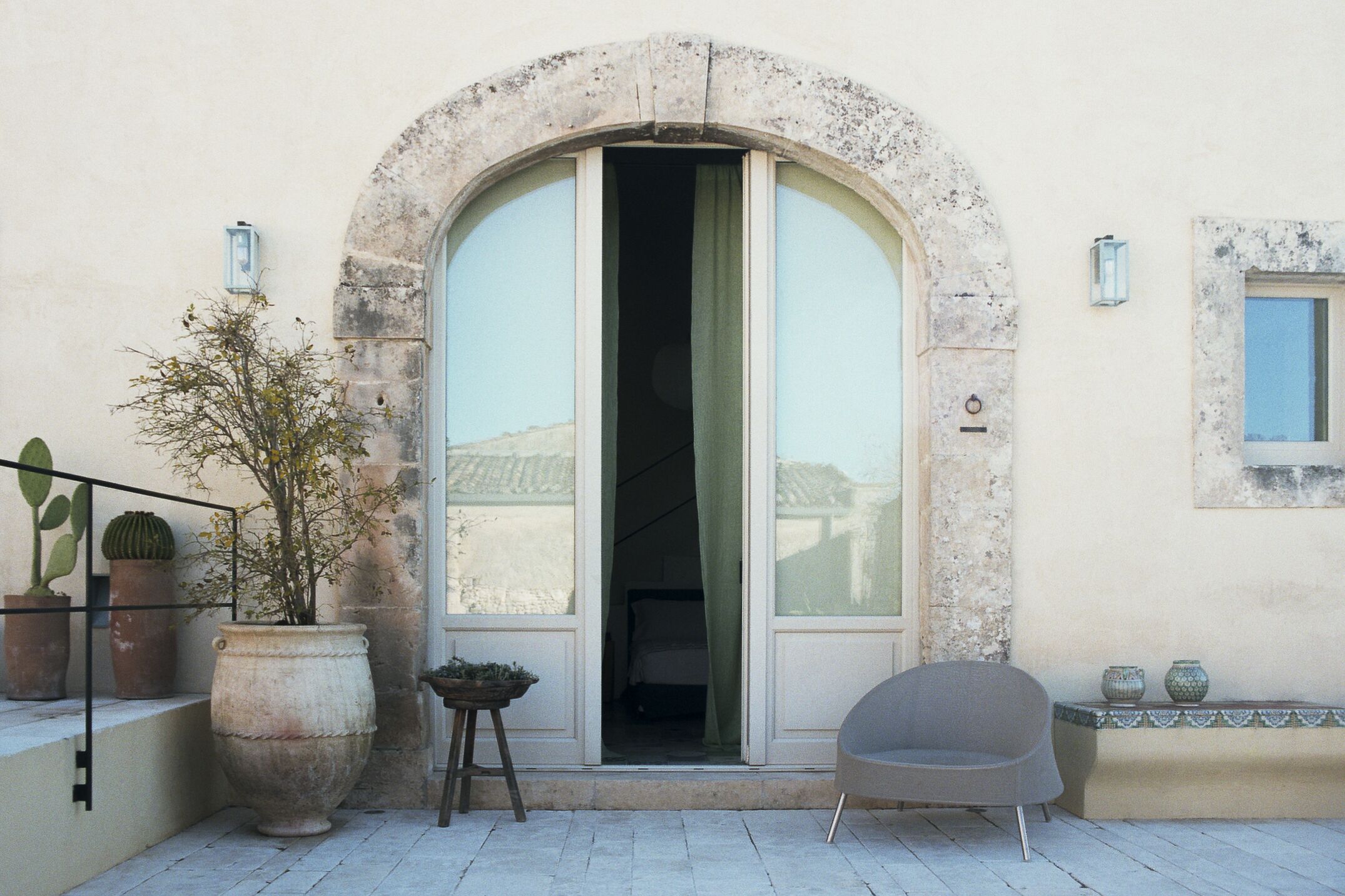 Une entrée d’immeuble avec une grande porte en arc de cercle, un pot de fleurs, une chaise et une petite table.
