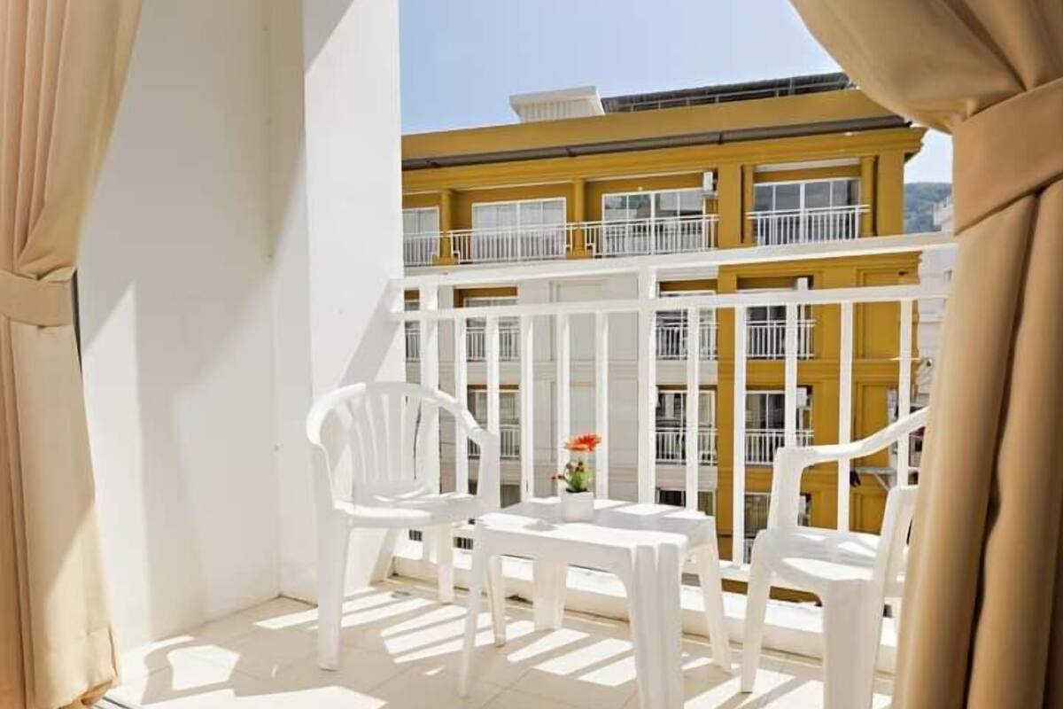 A balcony with white plastic chairs and a small table, overlooking a yellow building with white railings.
