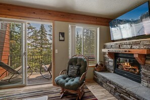 A cozy living room with a stone fireplace, a comfortable chair, and a large TV screen displaying a snowy landscape.