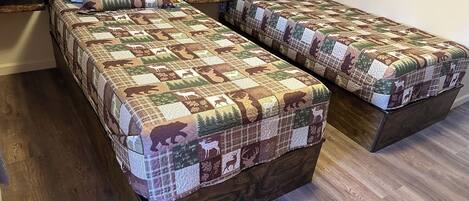 Duas camas de solteiro com colcha de patchwork estampada com padrões de animais, cabeceira escura e chão de madeira.