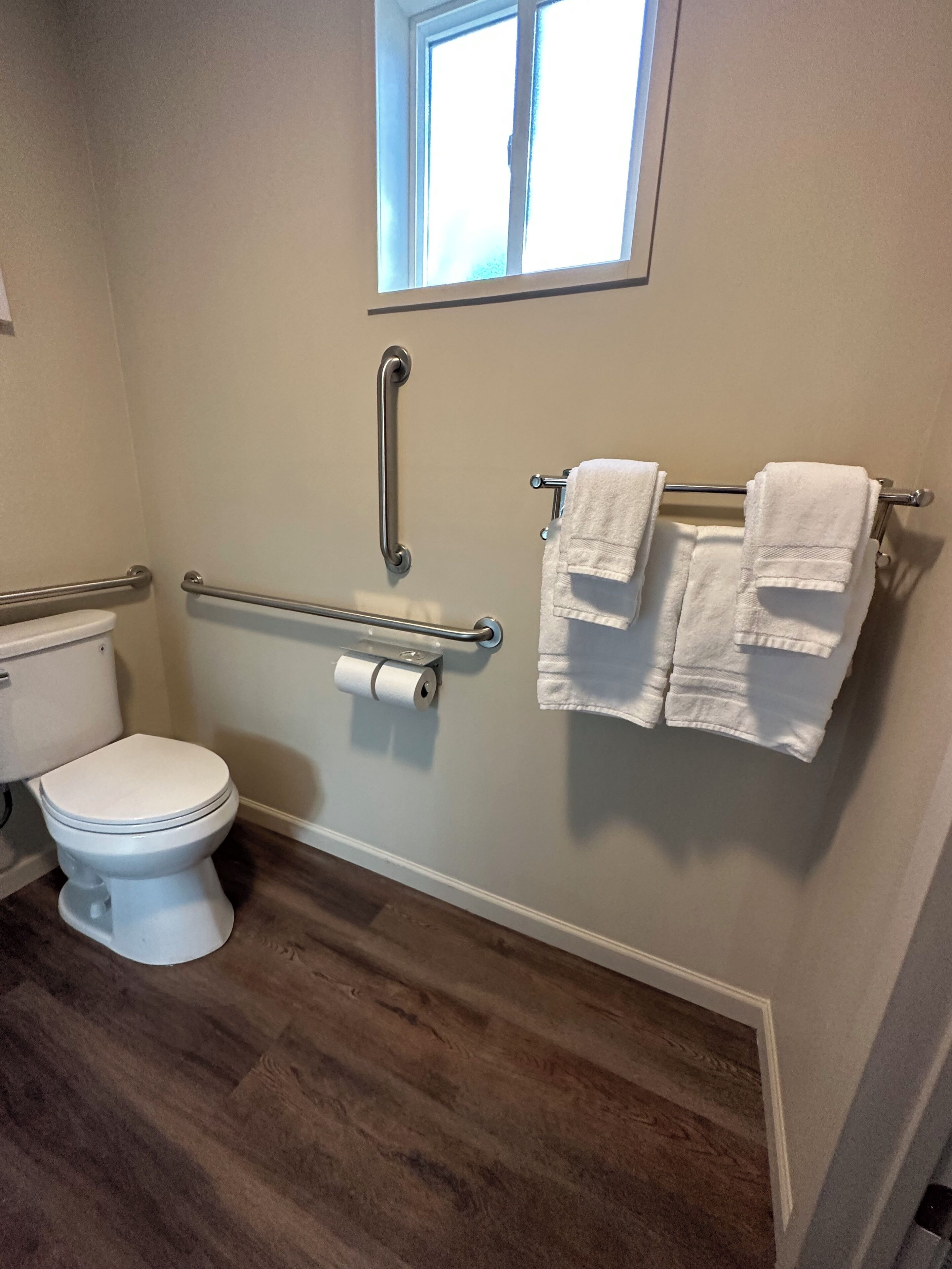 A bathroom with a white toilet, a handrail, a towel rack with two towels, and a window above the toilet.
