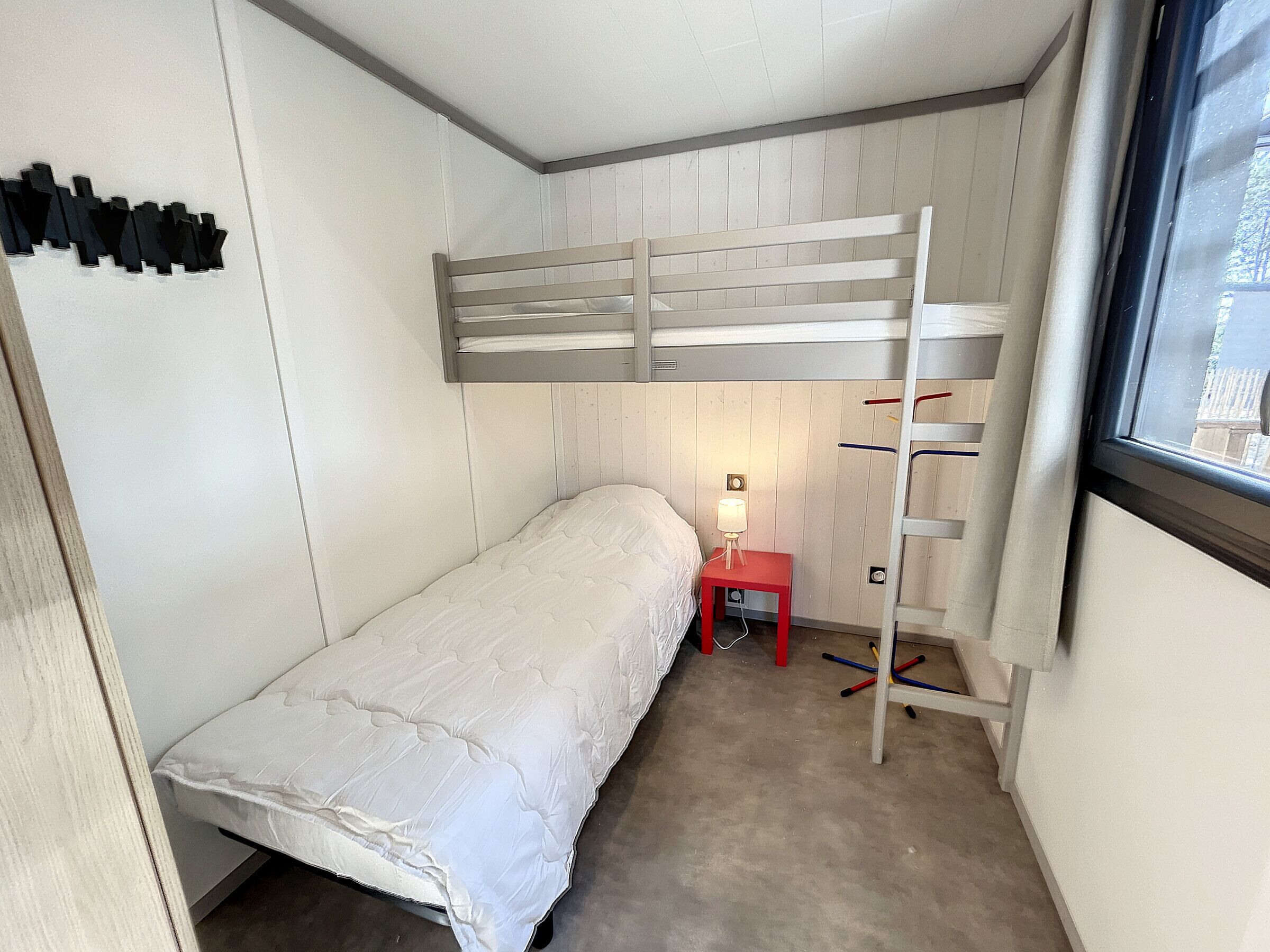 A small, single-bed room with a bunk bed, a bedside table with a lamp, a red chair, and a window with a view.