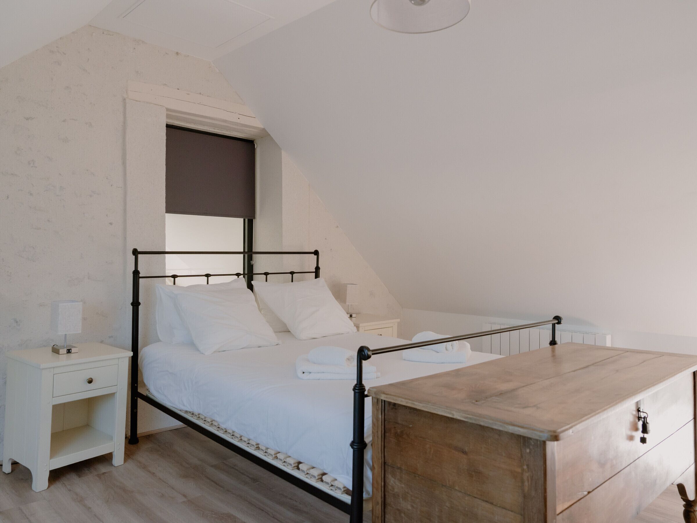 A bedroom with a sloped ceiling, a bed with white bedding, a wooden dresser, and a nightstand with a lamp.