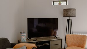 A living room with a flat-screen TV on a stand, a floor lamp, a sofa with cushions, and a small window with a plant.