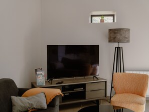Ein Wohnzimmer mit einem Flachbildfernseher auf einem Schrank, einer Stehlampe, einer Couch mit Kissen und einem kleinen Fenster mit einer Pflanze.