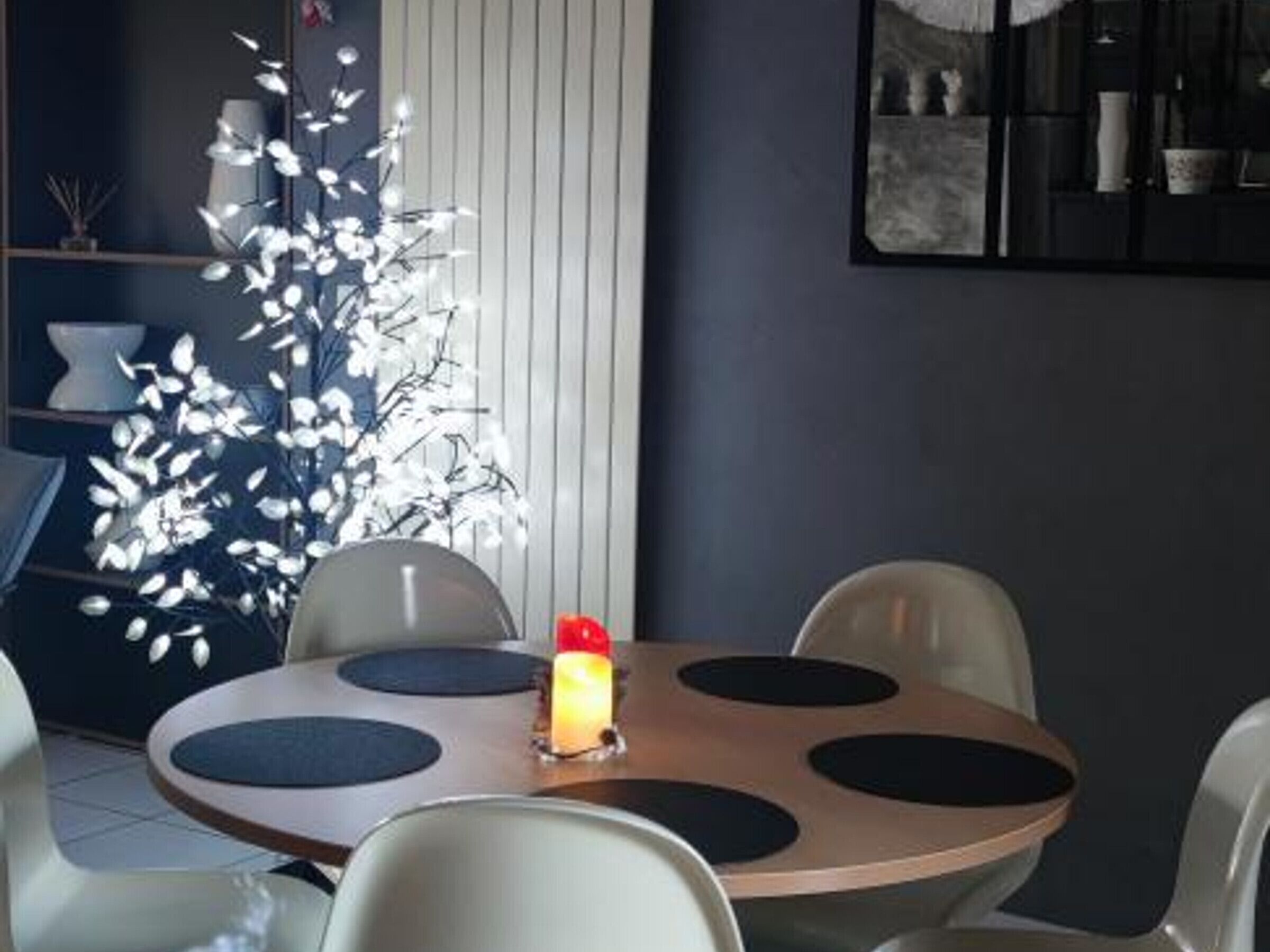 A dining area with a round table, white chairs, a decorative white tree, and a lit candle.