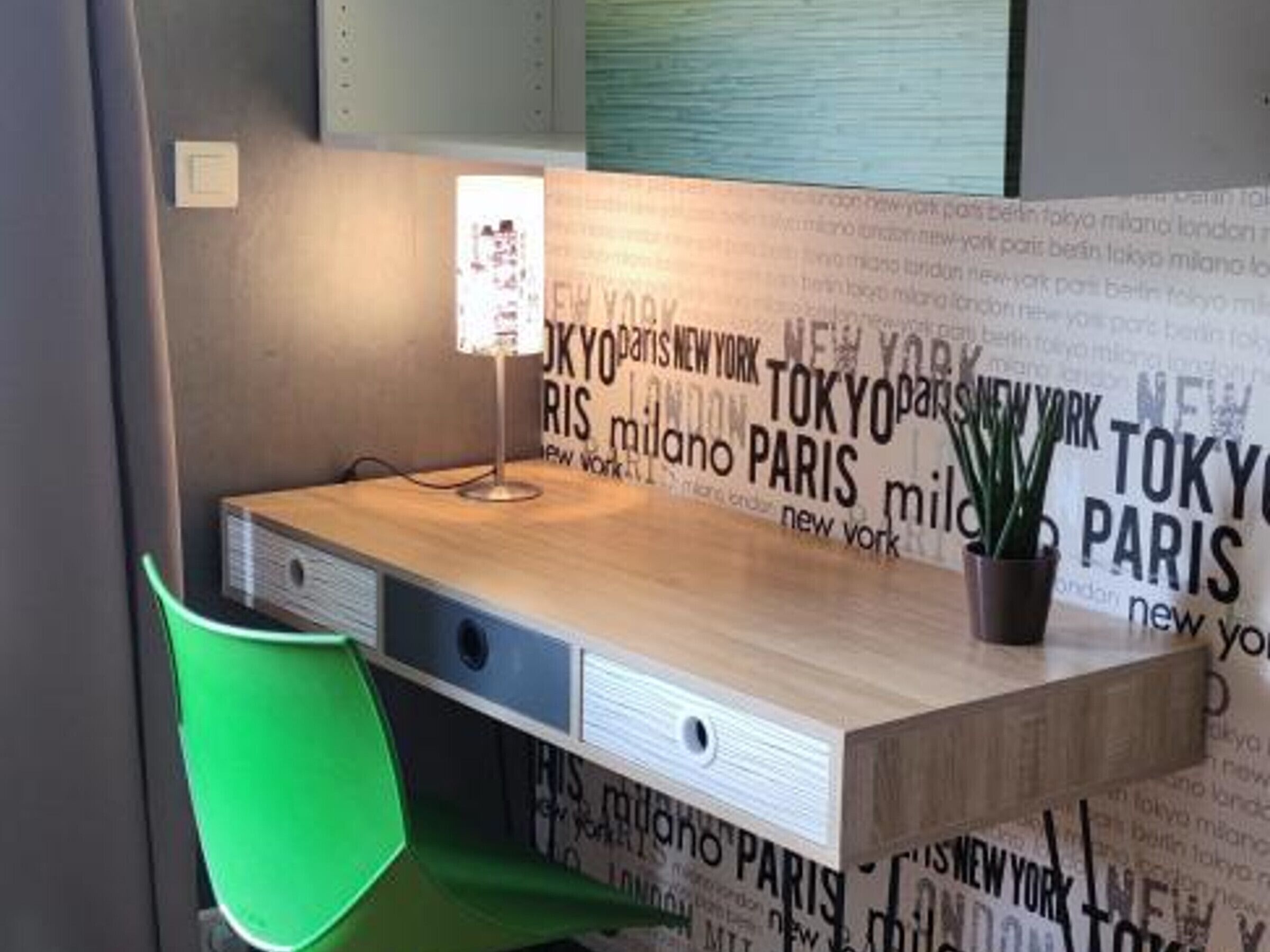 Un bureau avec une lampe, une chaise verte et un mur sur lequel sont écrits des noms de villes.