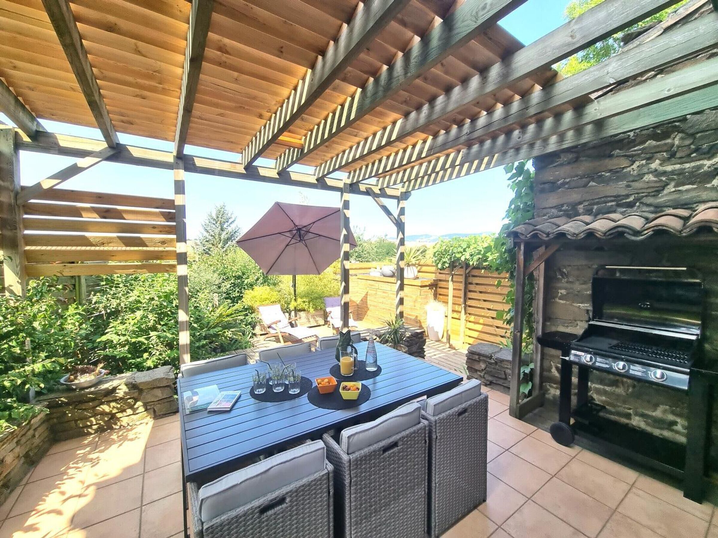 Un patio al aire libre con una pérgola de madera, una mesa con sillas, una parrilla y vistas al jardín.