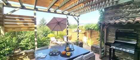 Un patio al aire libre con una pérgola de madera, una mesa con sillas, una parrilla y vistas al jardín.