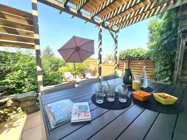 Une pergola en bois avec une table dressée pour les repas, des chaises d’extérieur, un grand parasol et une vue sur un jardin.