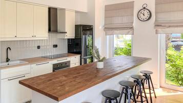 Une cuisine moderne avec des armoires blanches, un îlot central en bois, des tabourets de bar, un réfrigérateur en acier inoxydable et une horloge murale.
