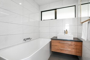 A modern bathroom with a white bathtub, a wooden vanity with a sink, and a large mirror above the vanity. - Redwoods Getaway - 100m to the Trails, Sleeps 7 (Rotorua)