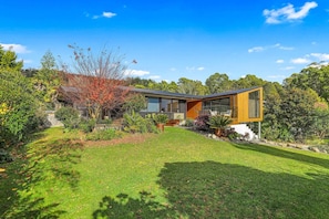 A modern house with a large glass facade, a well-maintained lawn, and a variety of plants and trees surrounding the property. - Redwoods Getaway - 100m to the Trails, Sleeps 7 (Rotorua)