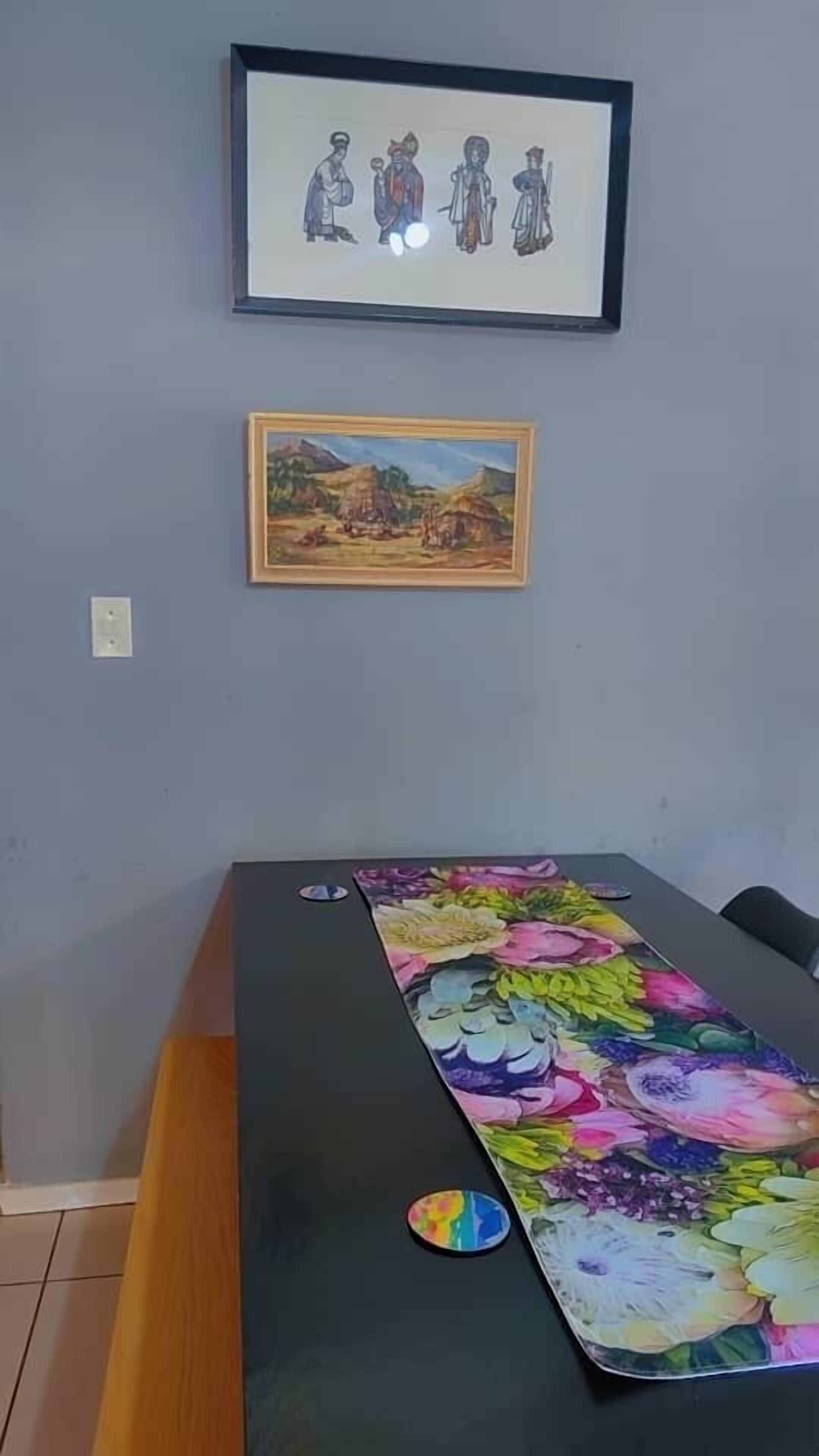 Une pièce avec une table recouverte d’une nappe à motifs floraux, un tableau encadré au-dessus de la table et une prise de courant sur le mur.
