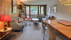 A cozy living room with a wooden wall, a bookshelf, a sofa, a dining table, and a large window offering a view of buildings and trees. - Large duplex for 8 people, south facing (La Plagne)