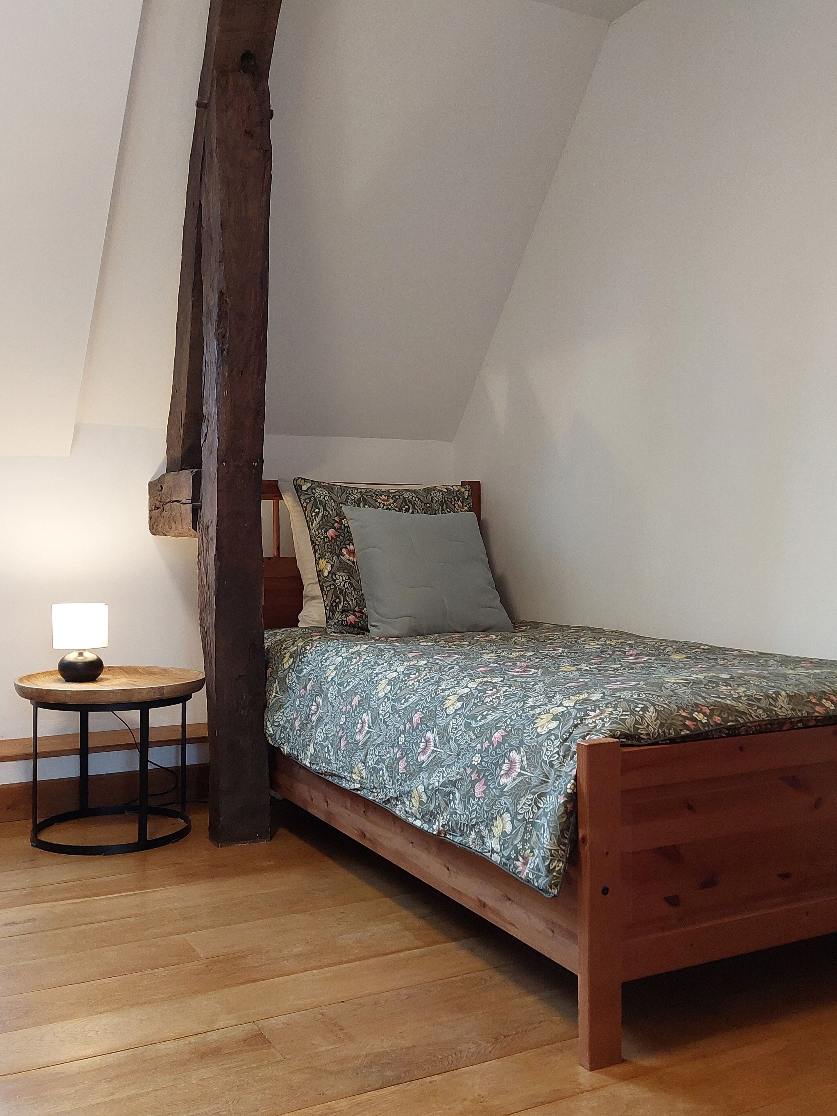 Un dormitorio pequeño y acogedor con una cama de madera, una mesita redonda con lámpara y un techo inclinado.