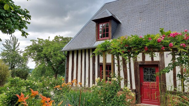 Una casa tradicional con techo inclinado, puerta roja y jardín con diversas plantas y flores.