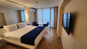 Quarto de hotel com uma cama grande, televisão, escrivaninha com cadeira e janela com cortinas.