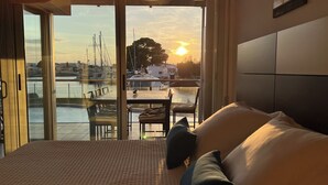 Ein Schlafzimmer mit einem großen Bett, Blick auf einen Yachthafen mit Booten sowie einem Essbereich mit Stühlen und einem Tisch.