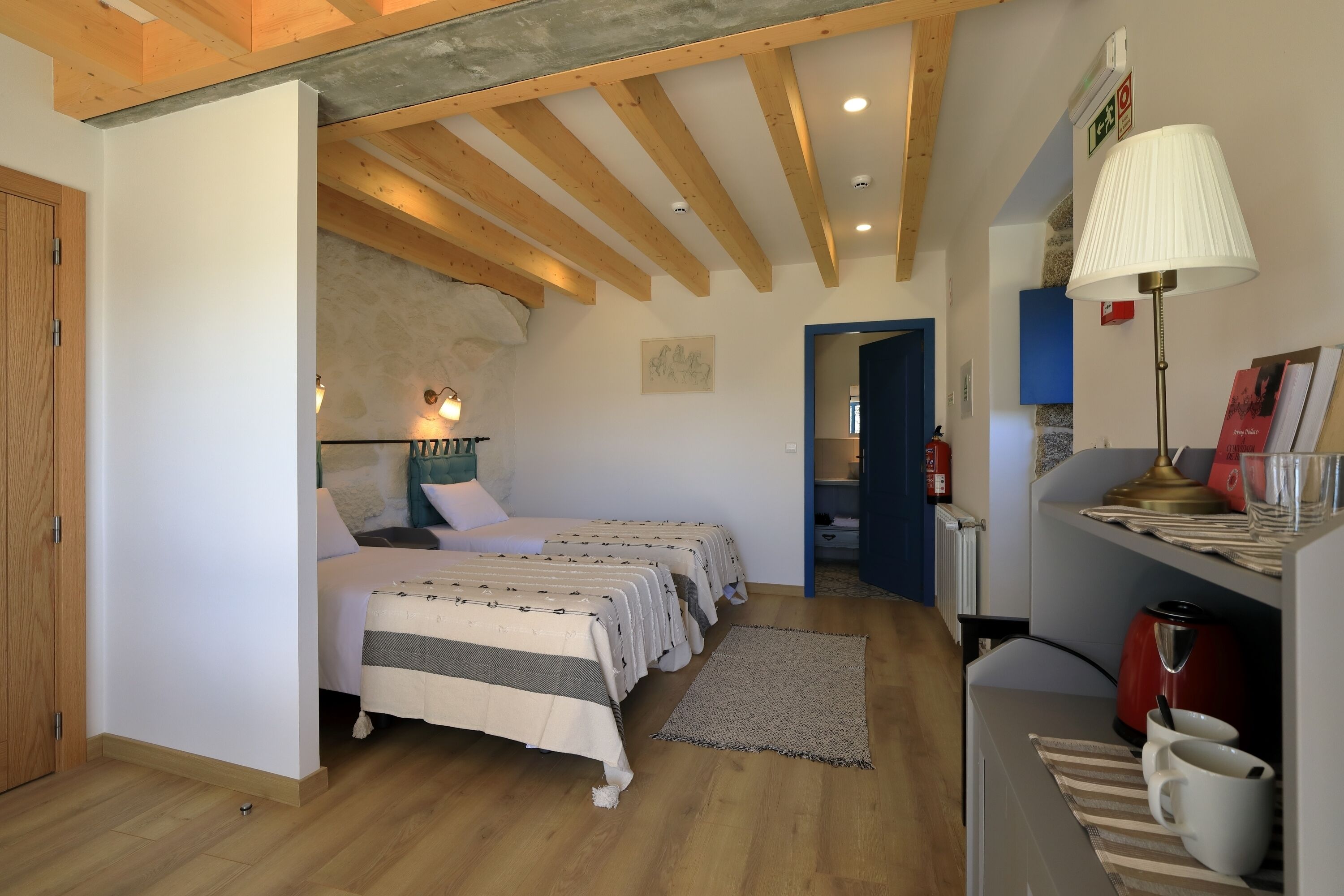 A bedroom with two beds, wooden beams, a lamp, a radiator, and a shelf with books and a kettle.