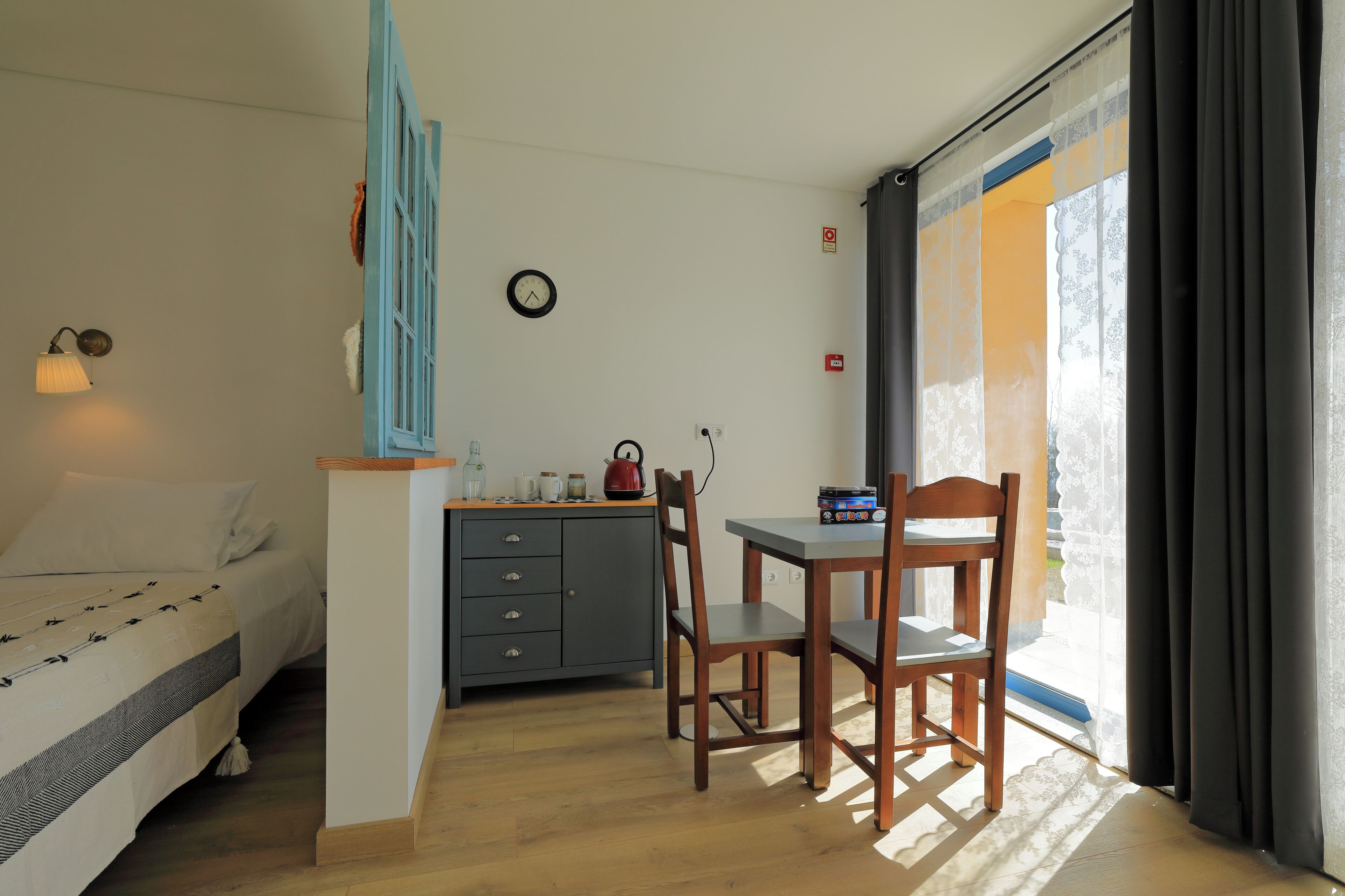 Un dormitorio con cama, escritorio con silla, una mesita con hervidor y ventana con cortinas.