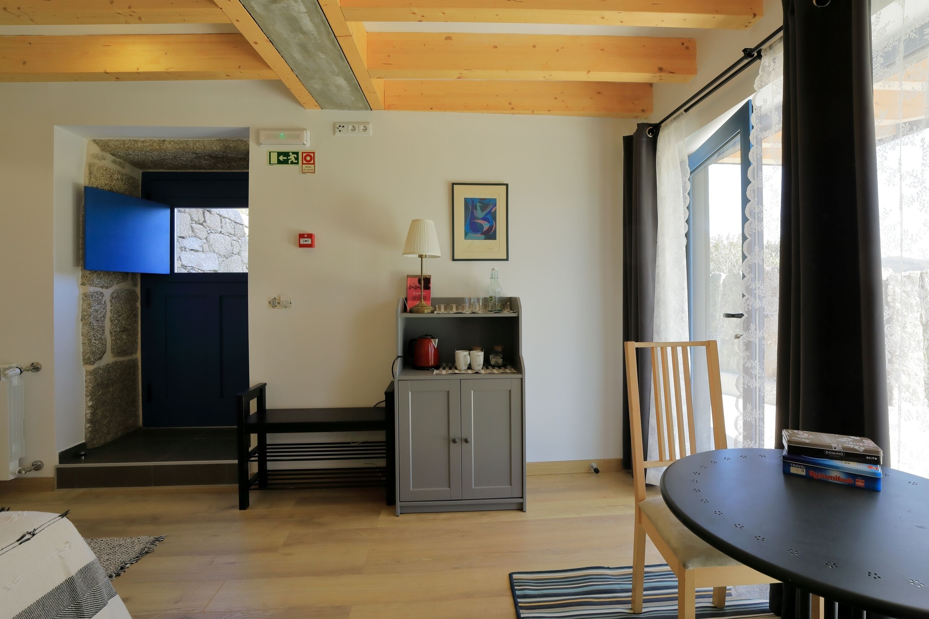 Una habitación con techo de madera, pared de piedra, puerta azul, armario gris, una mesa con libros y una silla.