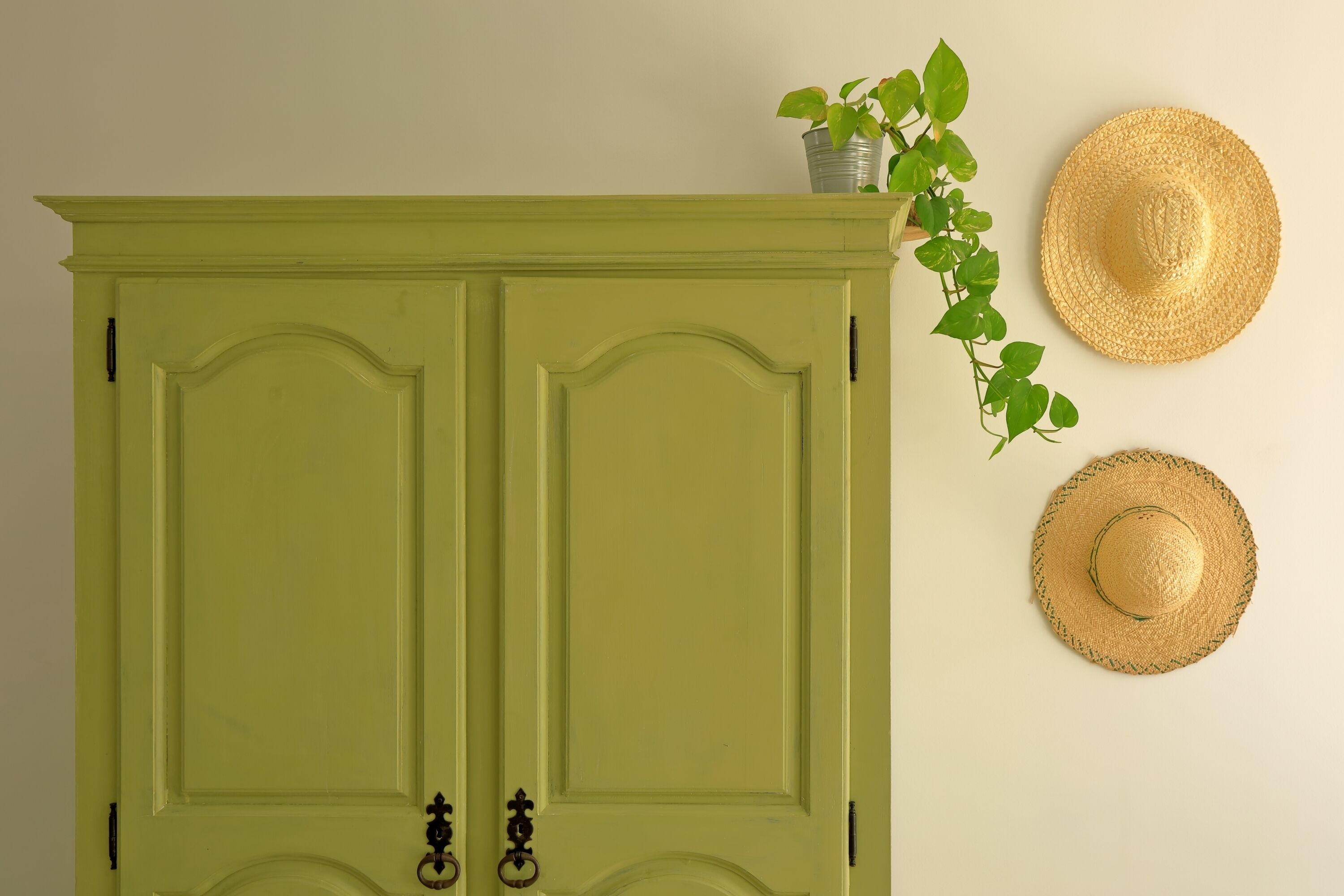 A green wardrobe with two doors, a potted plant on top, and two straw hats on the wall.