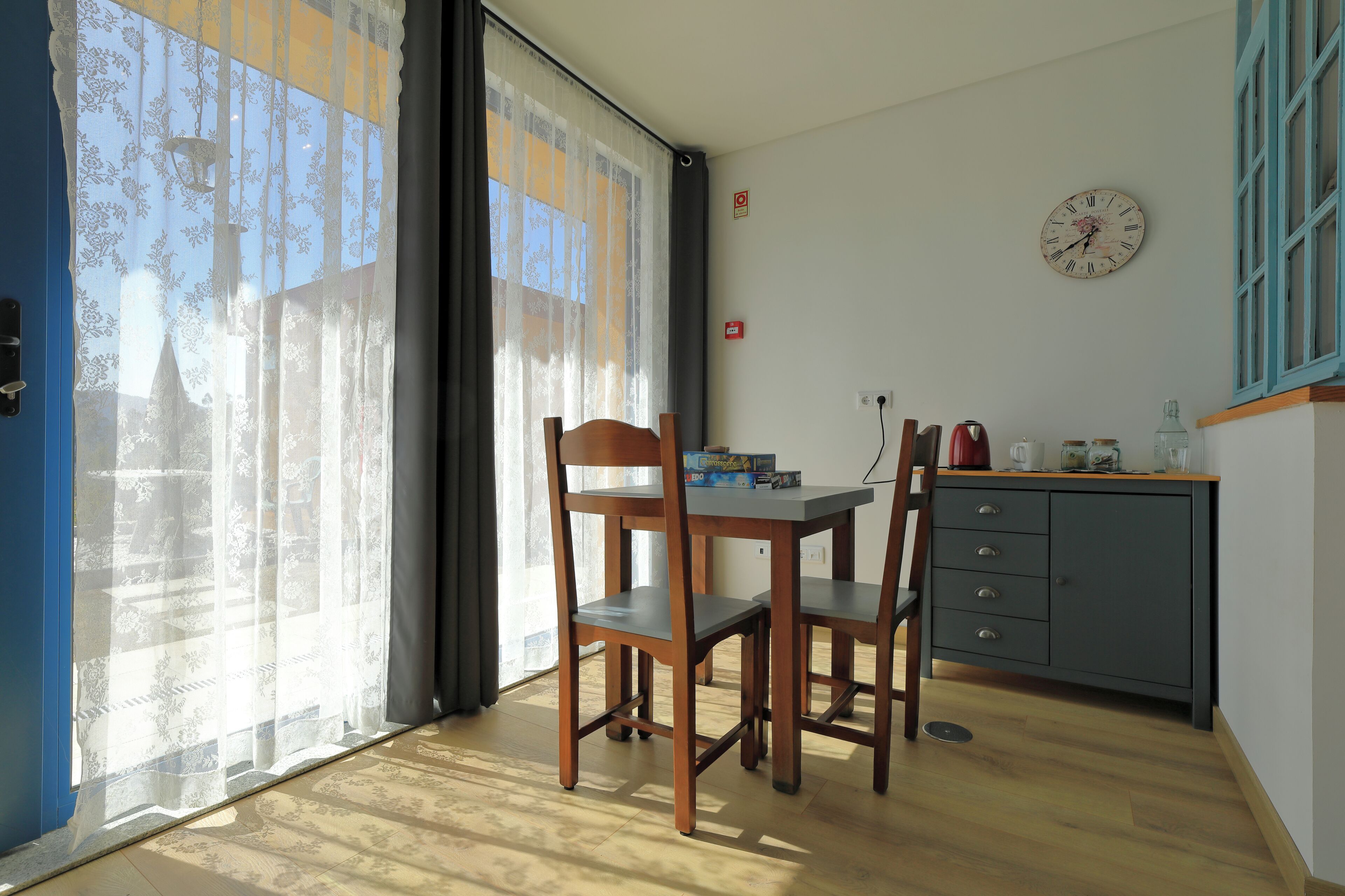A room with a dining table, chairs, a clock on the wall, and a window with sheer curtains.