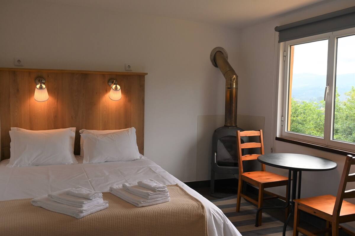 Ein Hotelzimmer mit einem Bett, zwei Nachttischlampen, einem Heizkörper, einem Tisch mit Stühlen und einem Fenster mit Blick.