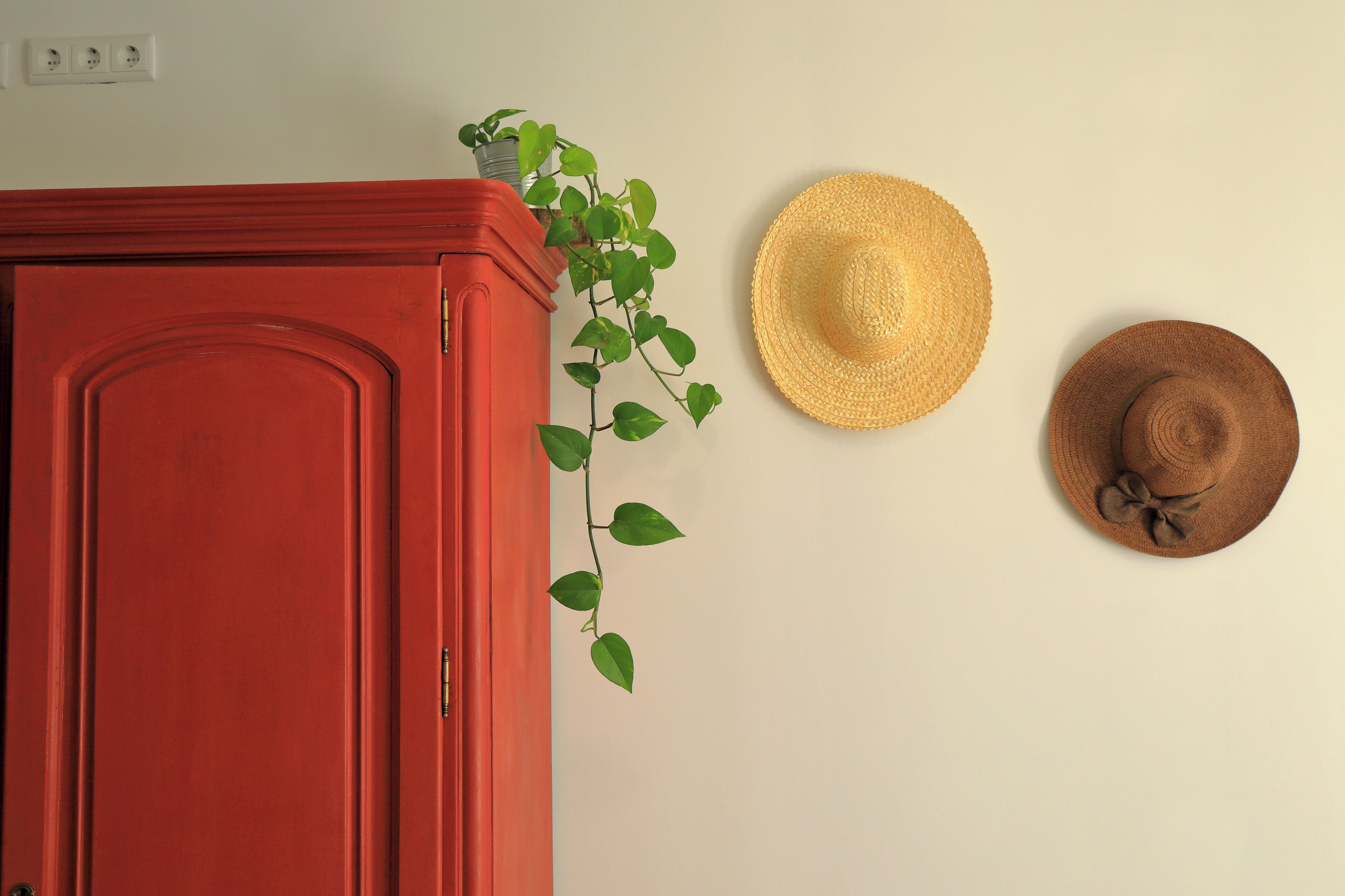 Un mueble rojo, una planta en maceta y dos sombreros en una pared.