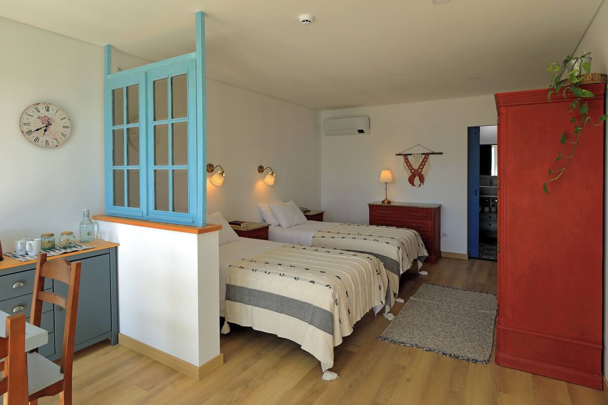 Ein Schlafzimmer mit zwei Betten, einem Schreibtisch und einem Stuhl. An der Wand hängt eine Uhr und über dem Bett befindet sich eine Klimaanlage.