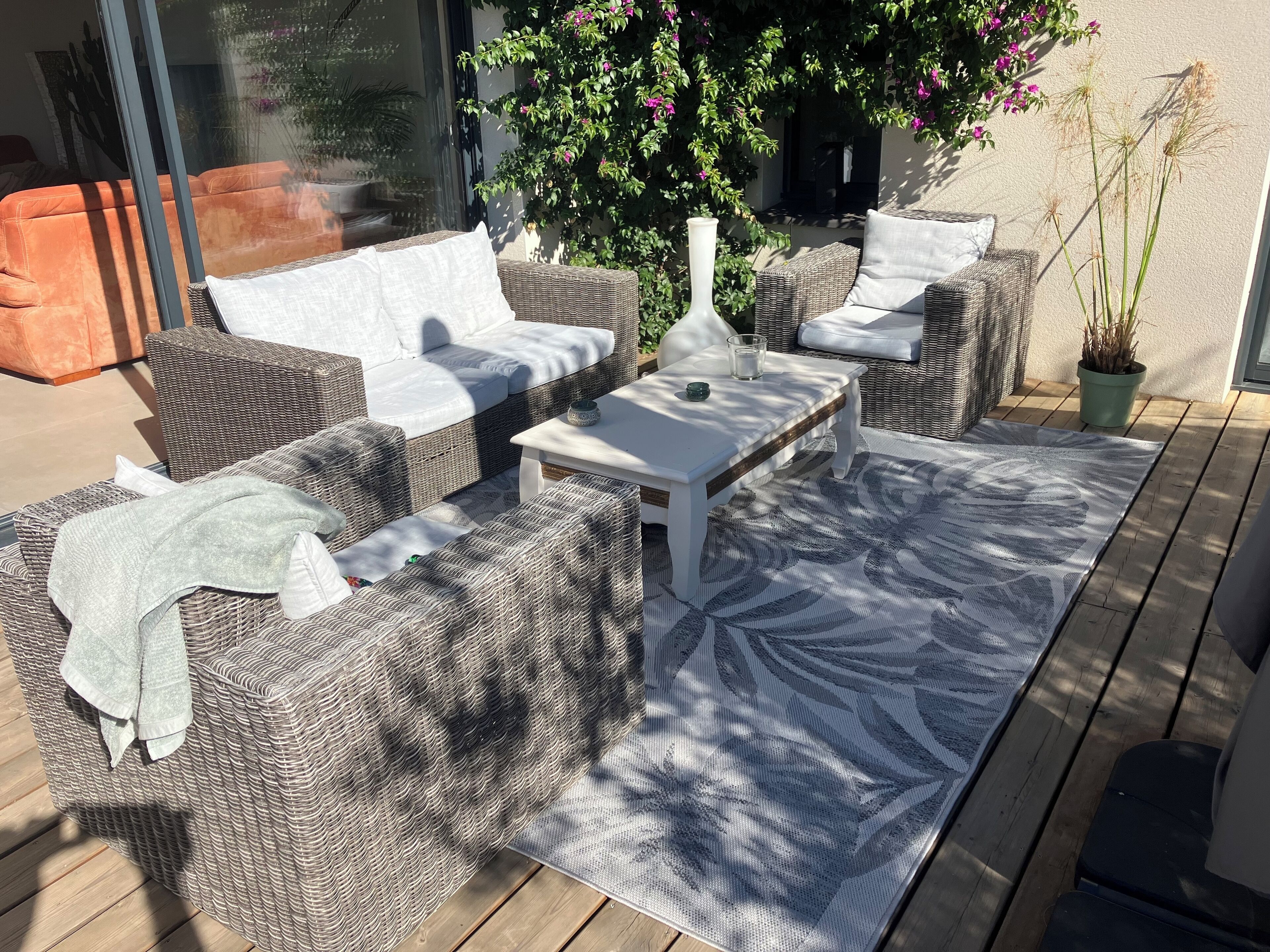 A sunlit outdoor seating area with wicker furniture, a white table, and a patterned rug.