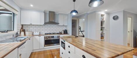 A modern kitchen with white cabinetry, a wooden countertop, and stainless steel appliances. There are two pendant lights, a window, and a door leading to another room.