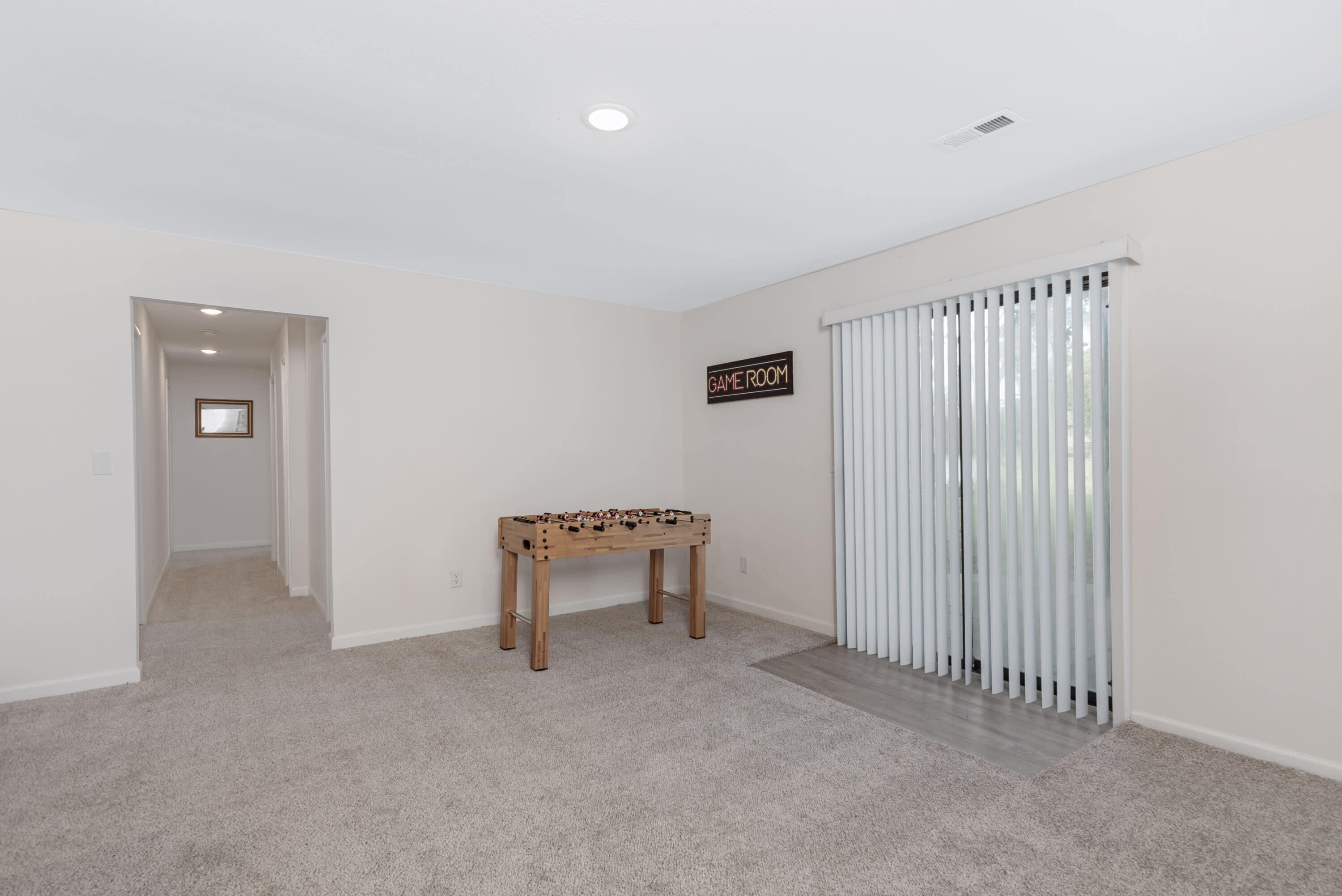 A room with a carpeted floor, a wooden foosball table, a sign labeled "GAME ROOM," and a window with vertical blinds.
