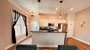 A modern kitchen with white cabinets, a central island, and pendant lighting. There are two green upholstered chairs and a glass table.