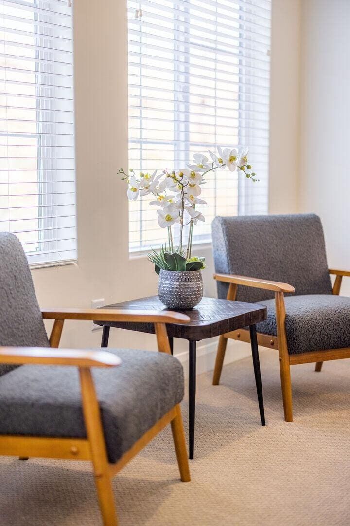 Ein Zimmer mit zwei grauen Polsterstühlen, einem kleinen Holztisch mit einer Orchidee im Topf und einem Fenster mit Jalousien.