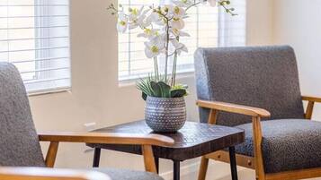 Ein Zimmer mit zwei grauen Polsterstühlen, einem kleinen Holztisch mit einer Orchidee im Topf und einem Fenster mit Jalousien.