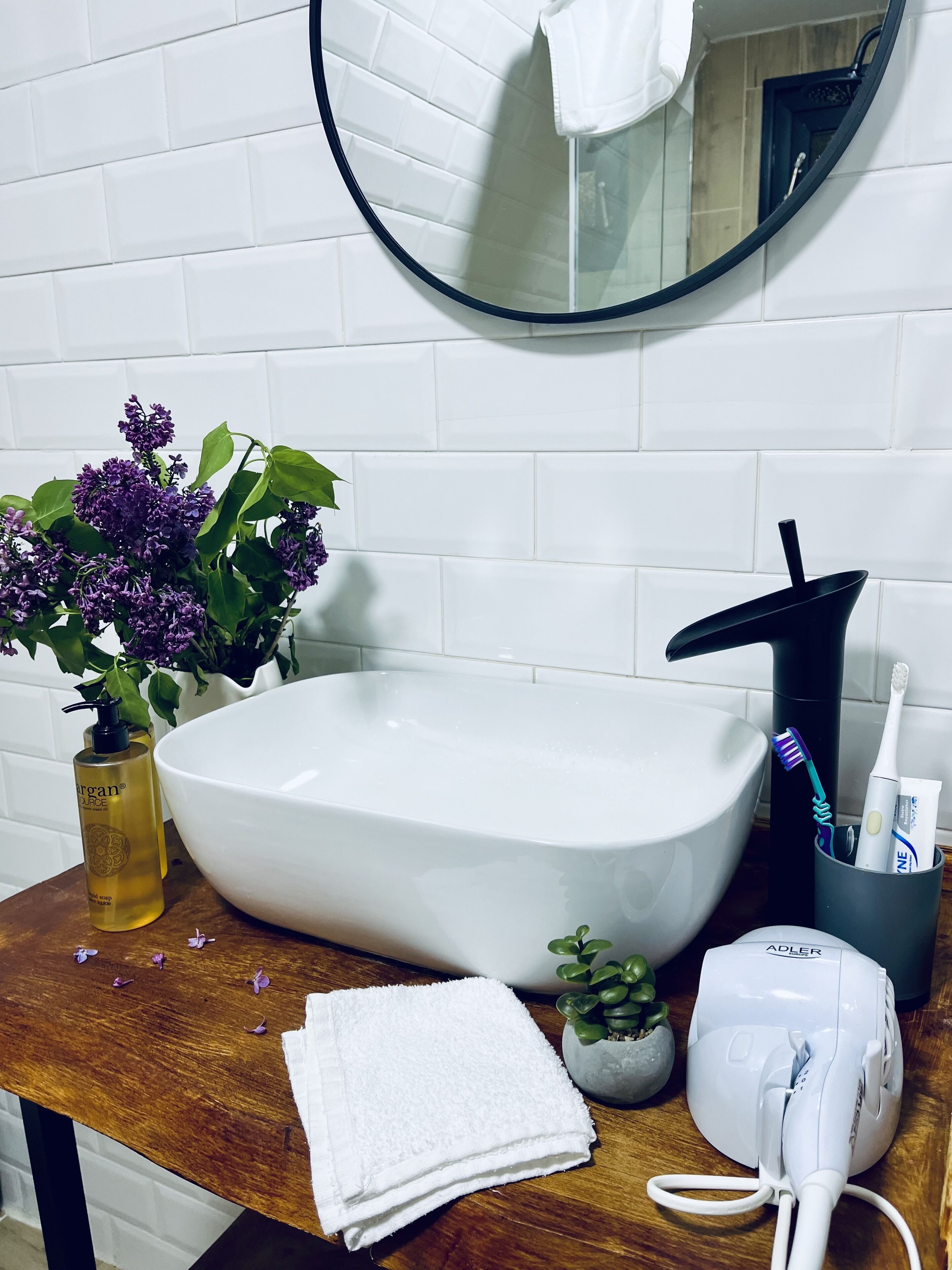 A bathroom with a white sink, a mirror, a potted plant with purple flowers, a bottle of liquid soap, a towel, and a hairdryer.