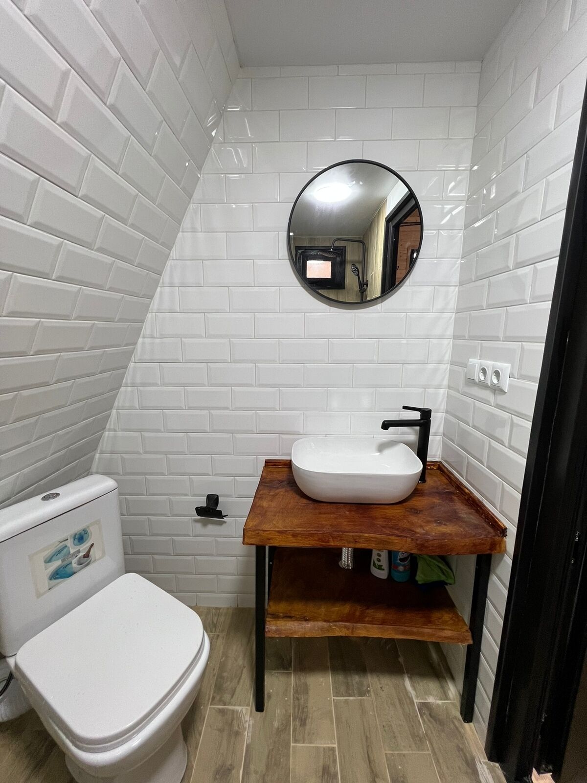 Un baño pequeño con paredes de azulejos blancos, un inodoro blanco, un tocador de madera con un lavabo y un espejo redondo.