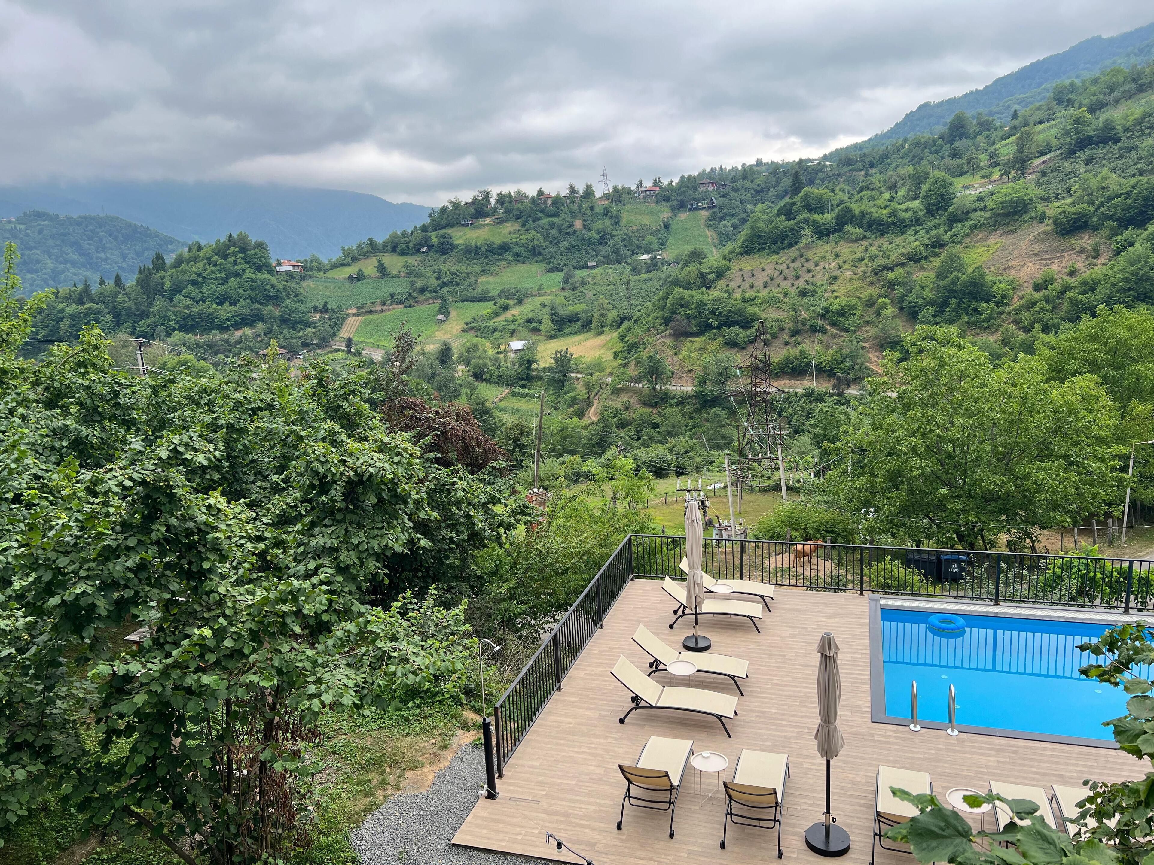 A pool area with lounge chairs and umbrellas, surrounded by lush greenery and hills.