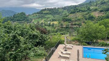 A pool area with lounge chairs and umbrellas, surrounded by lush greenery and hills.