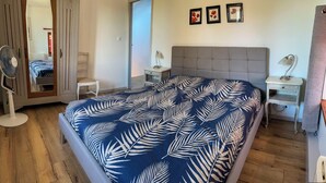Quarto com cama grande, piso de madeira, mesa de cabeceira com abajur, uma cadeira e um espelho.
